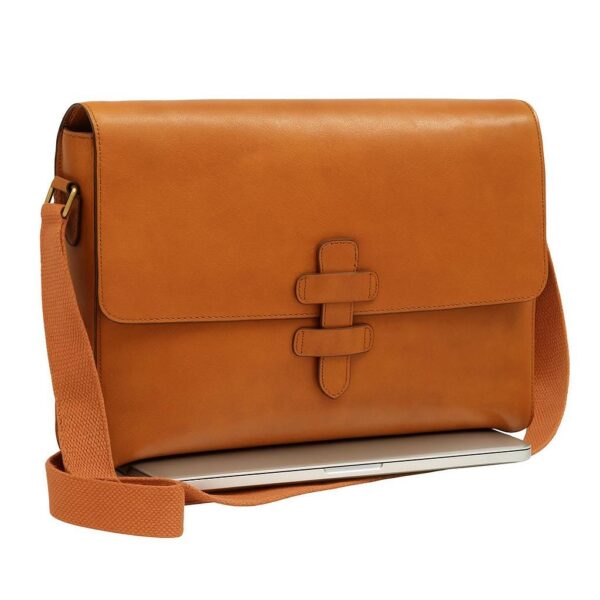 A tan, horizontal leather messenger bag with a large front flap and a vertical strap closure, resting on top of a closed silver laptop. The bag features a matching orange canvas shoulder strap.