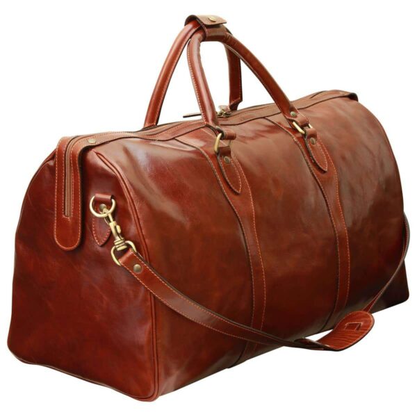 A large, **chestnut-brown leather duffel bag** shown from a three-quarter angle, featuring a rich, polished patina. It includes two rounded top handles with a snap-closure connector and a **detachable leather shoulder strap** with a sliding comfort pad. The bag is accented with **antique brass-toned hardware**, including swivel lobster clasps and side D-rings, and features a wide top-zipper closure that extends down the sides for easy access.