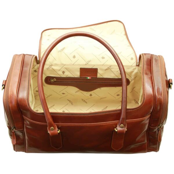An open cognac-brown leather duffel bag viewed from above, showing a cream-colored fabric lining with a repeat logo pattern, an internal zippered pocket with a leather brand patch, and large rounded leather carry handles.
