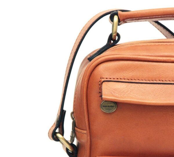 `Close-up detail of tan genuine leather bag showing Made in Italy brass badge, antique gold hardware and strap attachment, craftsmanship detail shot`