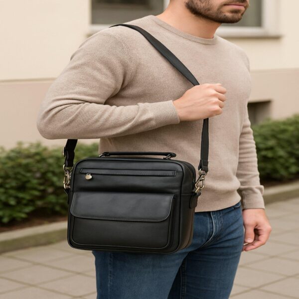 `Man in beige sweater and jeans wearing a black genuine leather crossbody messenger bag with front zip pocket and antique silver hardware, men's casual lifestyle shot outdoors`