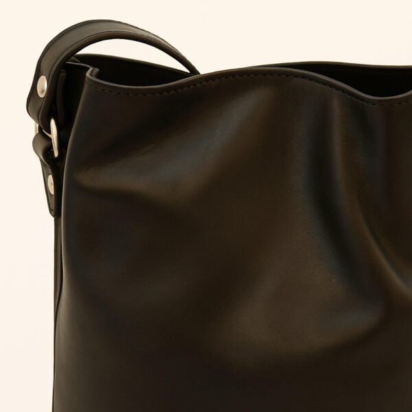 A close-up studio shot focusing on the top edge and strap attachment of a smooth black leather bag, featuring silver-toned rivets and clean stitching against a white background.