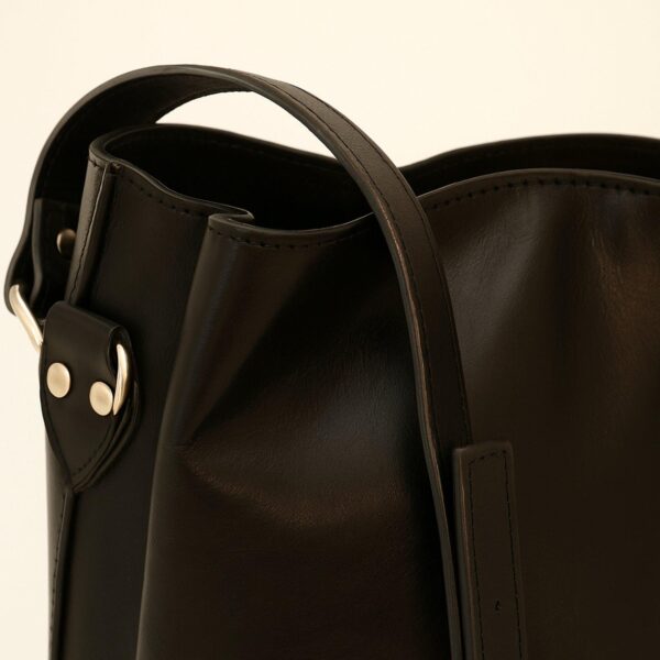 A close-up studio shot of a black leather bag's side and strap, highlighting the silver-toned rivets, a metal ring attachment, and the smooth, matte finish of the leather against a light background.