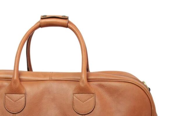 A close-up shot of the top section of a light brown leather briefcase, highlighting the sturdy rolled handles with a leather wrap connector and the reinforced stitching where the handles meet the bag's body.