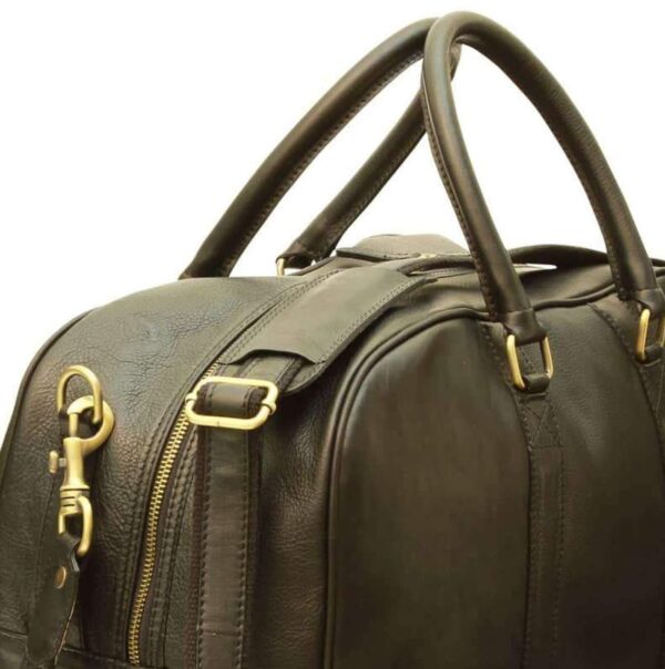 A close-up view of the top and side of a dark brown or black leather duffel bag. It features sturdy, rounded leather top handles attached with gold-toned rectangular rings. The bag also includes a detachable shoulder strap with a sliding leather comfort pad and a heavy-duty brass-toned lobster claw clasp. A high-quality metal zipper runs across the top, and the bag is shown against a plain white background.