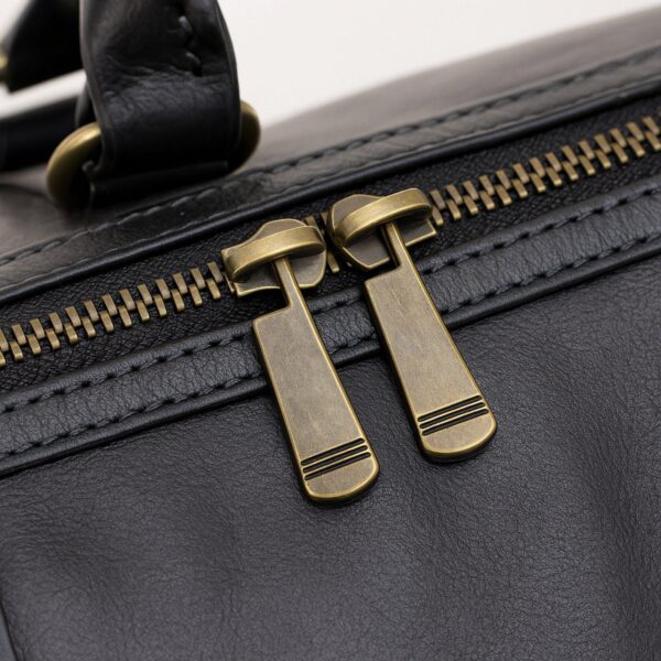 A close-up view of a black leather bag's main compartment, featuring a double-zipper closure with large, antique-brass-tone metal pull tabs and detailed parallel stitching along the zipper track.