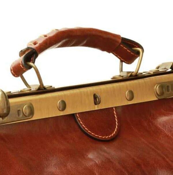 A close-up shot of a vintage-style brown leather doctor's bag, featuring a sturdy top handle with detailed stitching and a brass-toned metal frame with round rivets.