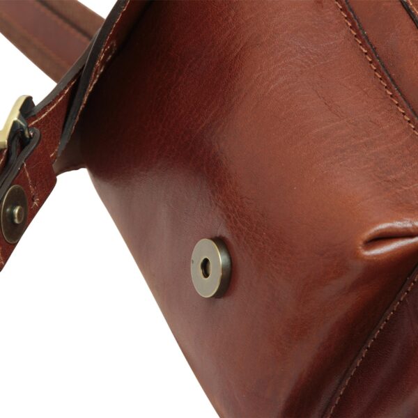A close-up shot of a brown leather bag, focusing on a magnetic snap fastener and the rich, pebbled texture of the leather. The image also shows a portion of a leather strap with a brass buckle and reinforced stitching along the bag's edges.