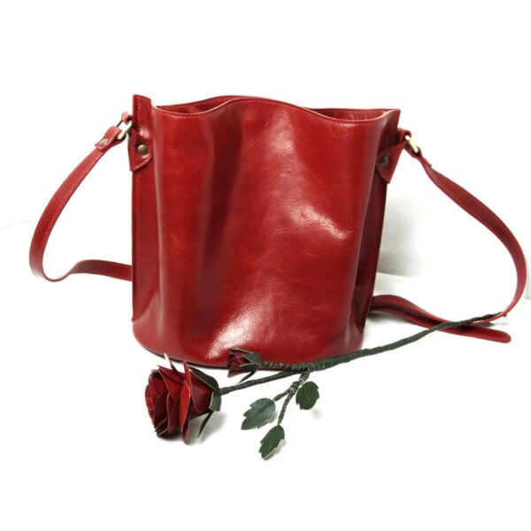 A red leather bucket bag with a matching shoulder strap sits on a white surface next to a handcrafted red leather rose with green leaves and a wire stem.