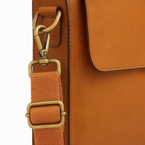 A close-up view of a tan leather messenger bag, highlighting the brass-tone metal swivel clasp and D-ring attachment for the adjustable orange canvas shoulder strap.