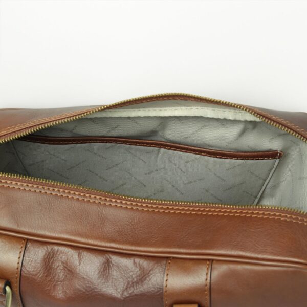 A top-down view into the open main compartment of a cognac leather briefcase, showing a grey patterned fabric lining with a leather-trimmed internal slip pocket and a brass-tone zipper track.