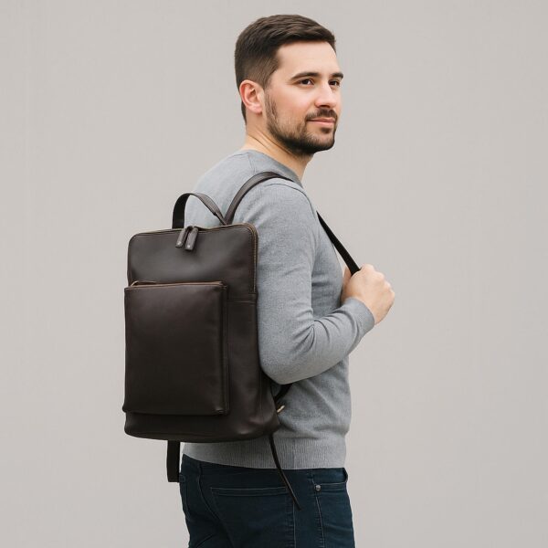 A man viewed from the side wearing a slim, dark brown leather backpack over a grey sweater and dark jeans, highlighting the bag's sleek profile and rectangular design.