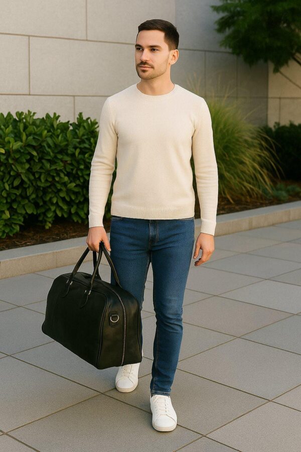 A man dressed in a cream crewneck sweater, blue slim-fit jeans, and white sneakers walks along a paved outdoor path while carrying a large black leather duffel bag.