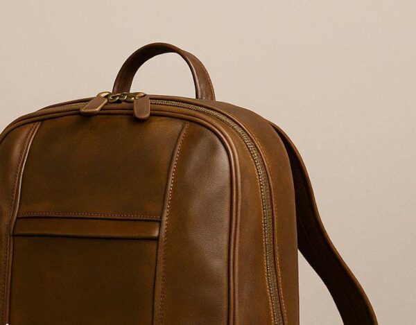A close-up, high-angle view of the top of a brown leather backpack. The image focuses on the curved top handle, dual brass zippers with leather pulls, and the fine, tonal stitching along the seams and front pocket.