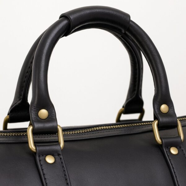 A close-up view of the black leather top handles of a duffel bag, featuring reinforced stitching, brass-tone rivets, and rectangular metal loops connecting to the main body.
