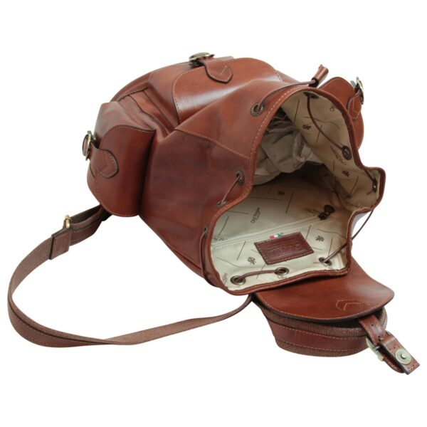 An open cognac-brown leather backpack lying on its side, revealing a cream-colored fabric lining patterned with logos, a zippered interior pocket with a leather brand patch, and a drawstring closure system with metal eyelets.
