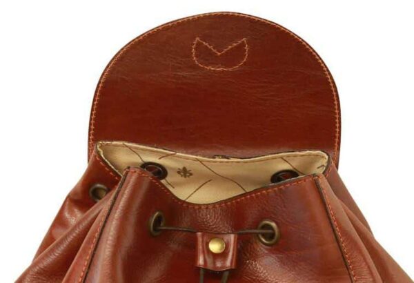 A close-up shot of the open top compartment of a vintage-style brown leather backpack, featuring a drawstring closure with brass-toned eyelets and a magnetic snap button on the leather flap. The interior is lined with a light-colored fabric patterned with small logos.