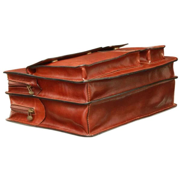 A low-angle side view of a cognac-colored leather briefcase, showing its multi-layered gusseted structure with two main compartments and side zippers with brass-tone pulls.