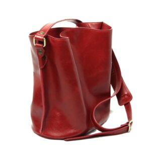 A side-angle view of a deep red, slouchy leather bucket bag against a white background. The image highlights the brass hardware, adjustable shoulder strap, and the soft, polished sheen of the leather