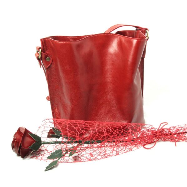 A deep red leather bucket bag stands behind a handcrafted leather rose. The rose is laid across a delicate red decorative mesh netting on a white background, highlighting the bag’s rich texture and the artistic presentation.